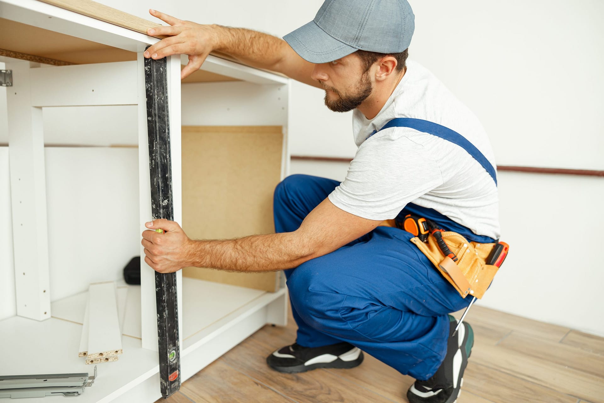 Professional handyman, male worker in uniform using spirit level while checking measurements of kitchen cabinet indoors